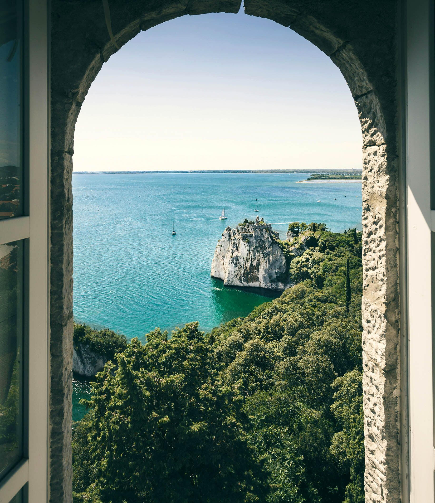 View through stone arch to turquoise water and forested cliffs — a quiet threshold, reflecting Terra Noma’s invitation to intentional exploration